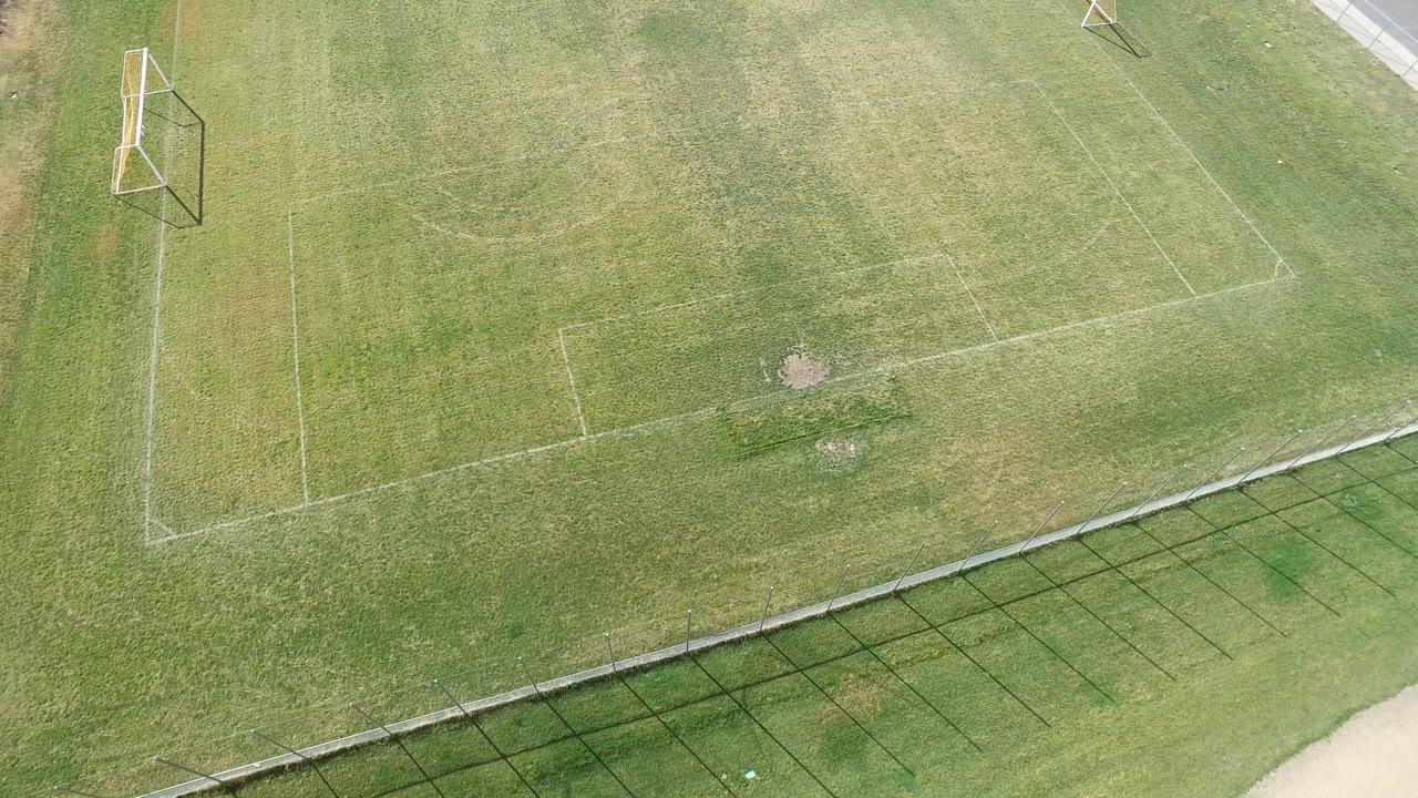 Overhead frame of a Hidden Lake, Colorado soccer field with a goal near the upper left, white boundary lines across green grass, fence shadows at the bottom, and no visible people.