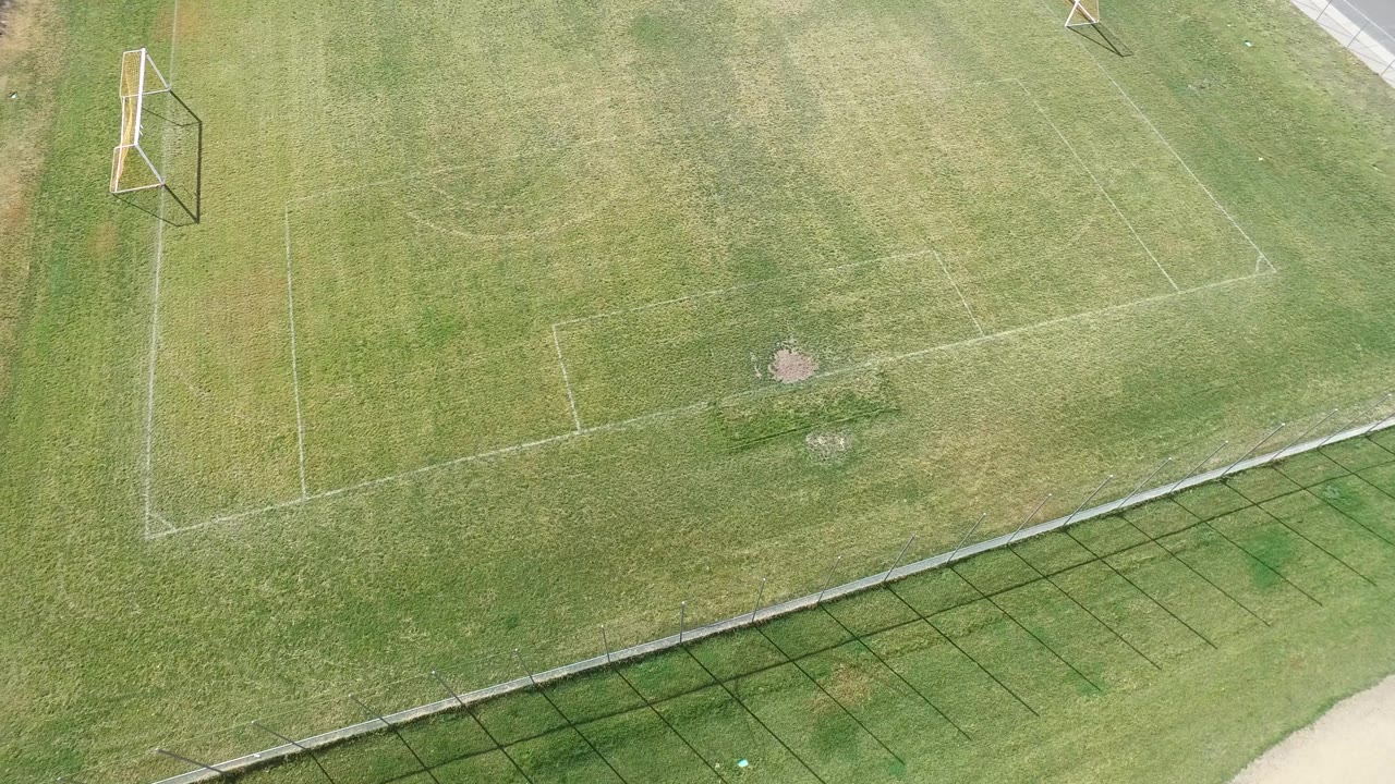Aerial frame over the Hidden Lake soccer field with the goal closer to the left edge, white markings on worn green turf, fence shadow along the lower edge, and no visible people.
