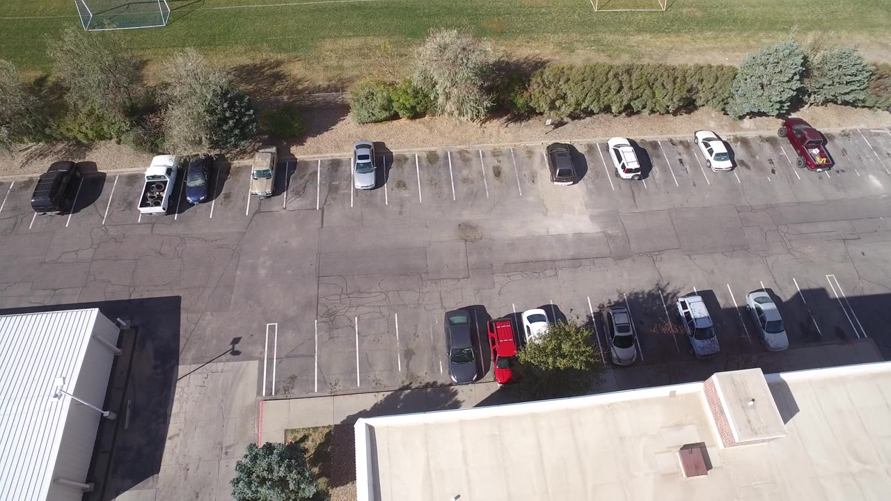 High overhead view of a Hidden Lake, Colorado commercial parking lot with parked cars, a flat-roof building at the bottom edge, grass field above, and no visible people.
