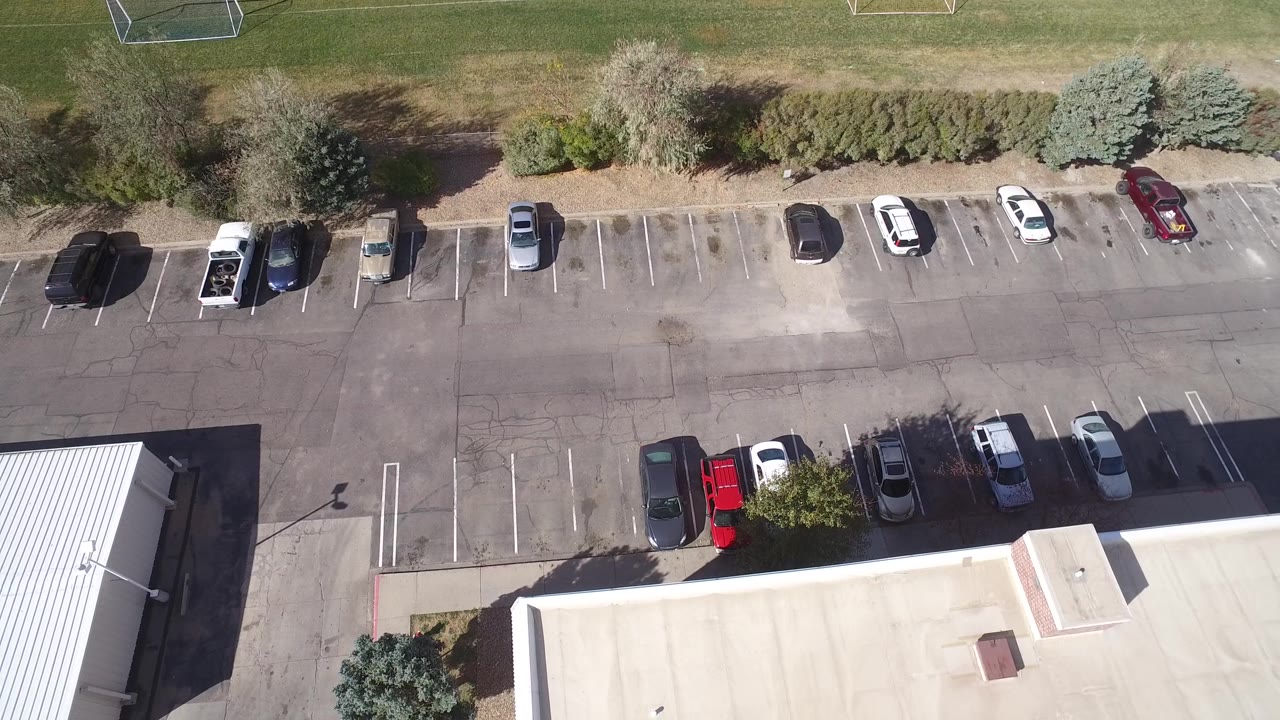 Overhead frame in Hidden Lake, Colorado showing rows of parked vehicles, landscaped shrubs, a pale commercial rooftop, open parking spaces, and no visible people.