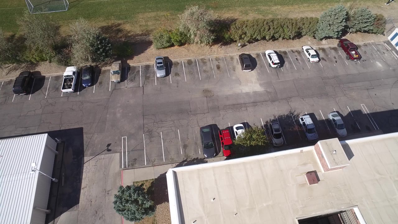 Top-down parking area with cars, building roof, trees, and field edge.