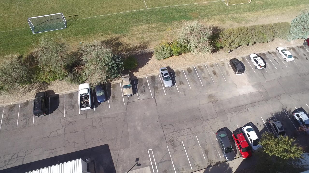 Aerial frame over Hidden Lake, Colorado showing a paved parking lot with scattered cars, trees, and a soccer goal near the upper field edge, with no visible people.