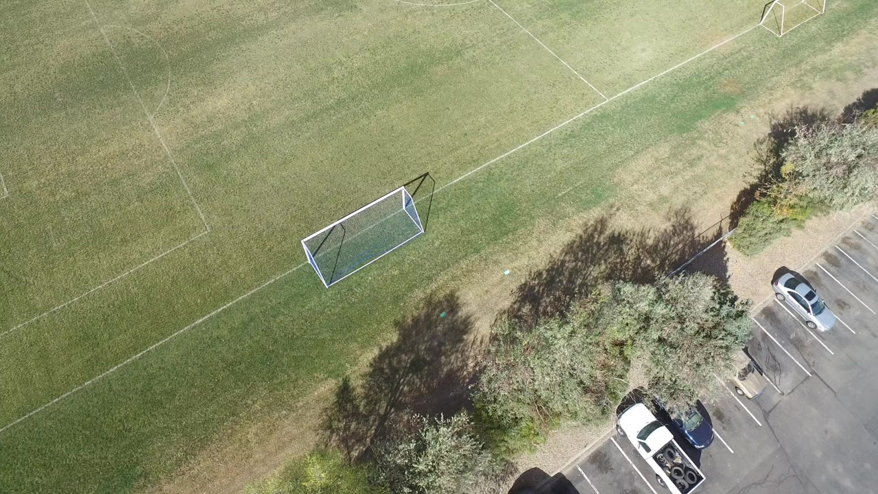 High overhead view of Hidden Lake, Colorado soccer fields at the start of the clip, with one goal on the grass, trees along the edge, parked cars, and no visible people.