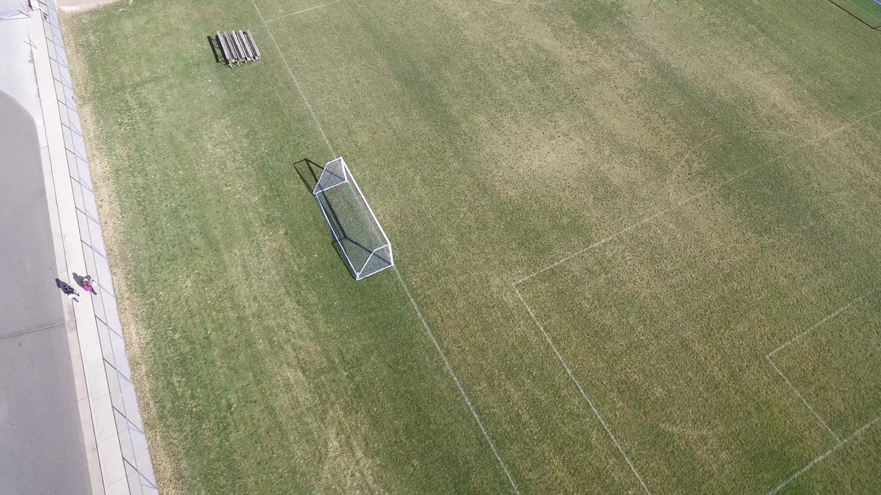 Aerial frame in Hidden Lake, Colorado with the soccer goal left of center, faint field lines across the grass, and two small people visible on the sidewalk near the road.