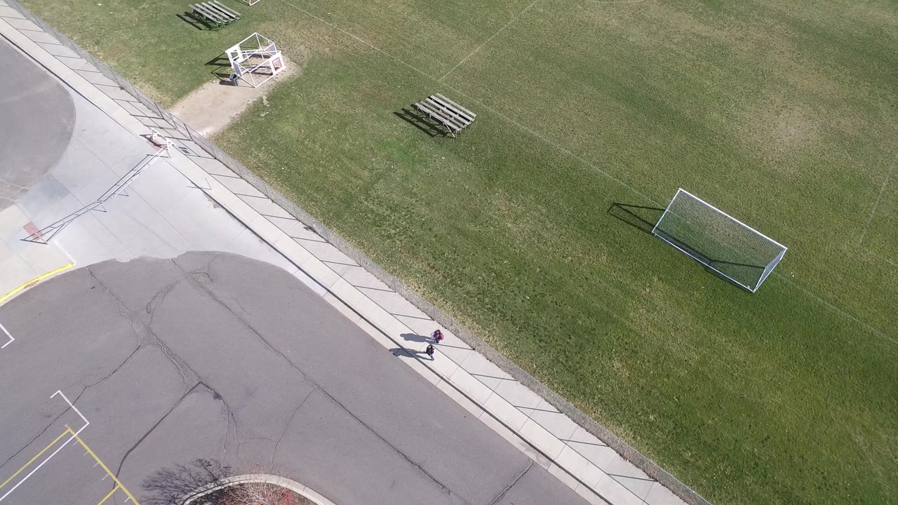 Aerial frame in Hidden Lake, Colorado with the sidewalk cutting diagonally between the paved road and open grass, plus benches, a small soccer goal, and a distant pedestrian.