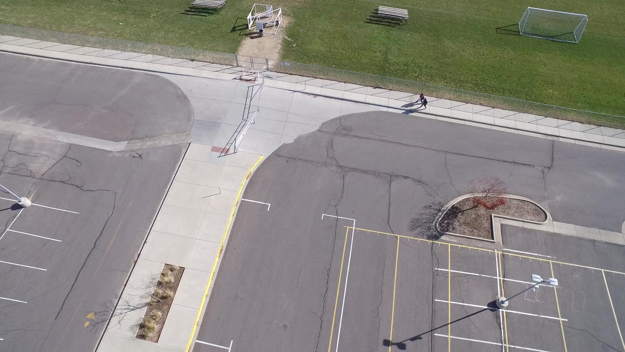 Drone view in Hidden Lake, Colorado, looking down at parking spaces, curbs, a grassy field with goals, and a small person continuing along the top sidewalk.