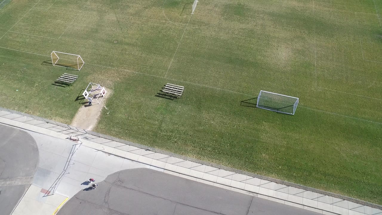 Drone view over the Hidden Lake, Colorado grass field with two soccer goals, bleacher rows, a small paved path area, and tiny distant people at the lower edge.
