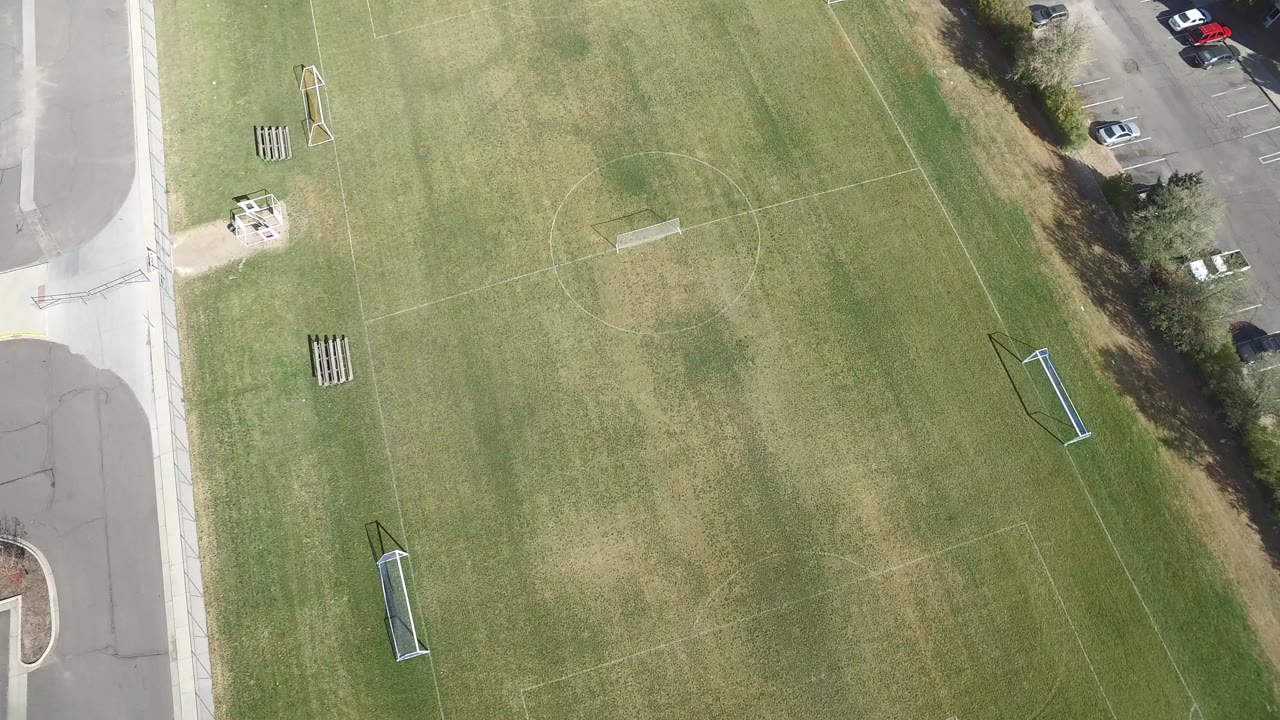 Overhead view of a grass sports field beside a road and parked cars.