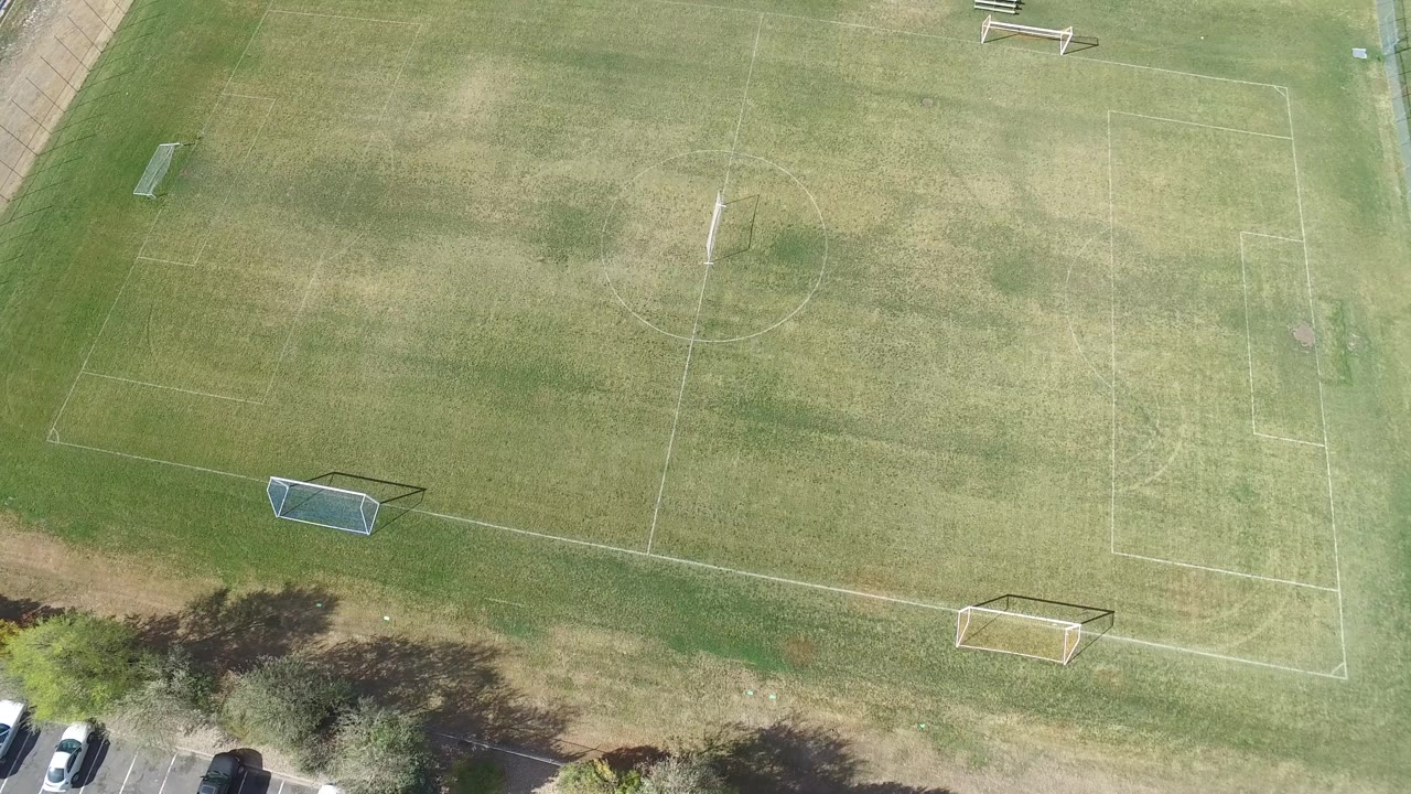 Top-down frame in Hidden Lake, Colorado showing a lightly worn grass field, goals, benches near the upper edge, a road corner, and no visible people.