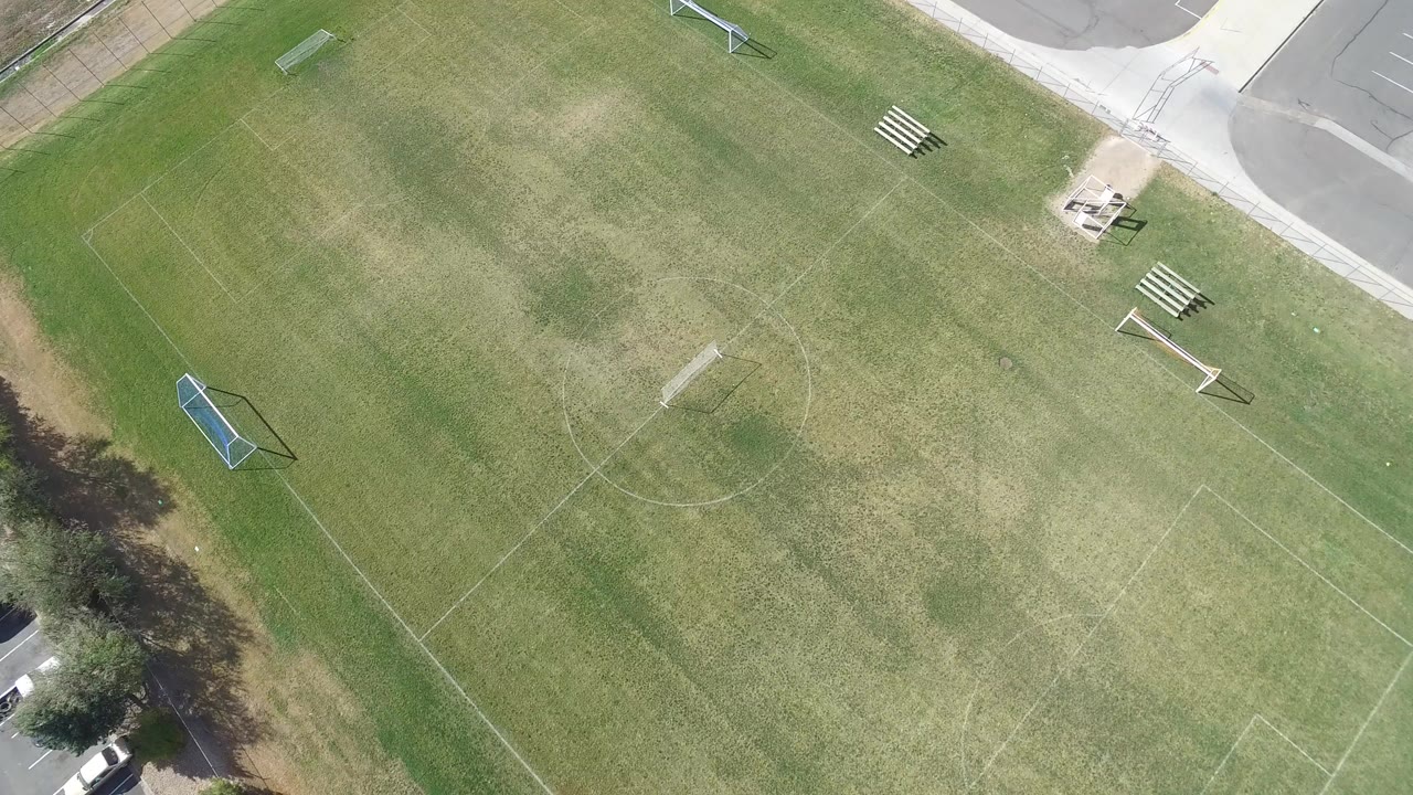 Overhead frame of a grass soccer field in Hidden Lake, Colorado, with faint field lines, a blue goal at left, sidewalk and pavement at the top right, trees along the lower edge, and no visible people.