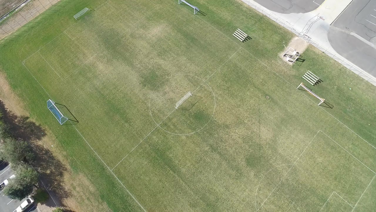 Bird's-eye frame in Hidden Lake, Colorado, showing the grass field shifted slightly in view, with a blue goal near the left side, bleachers near the upper right, sidewalk, pavement, trees, and no visible people.