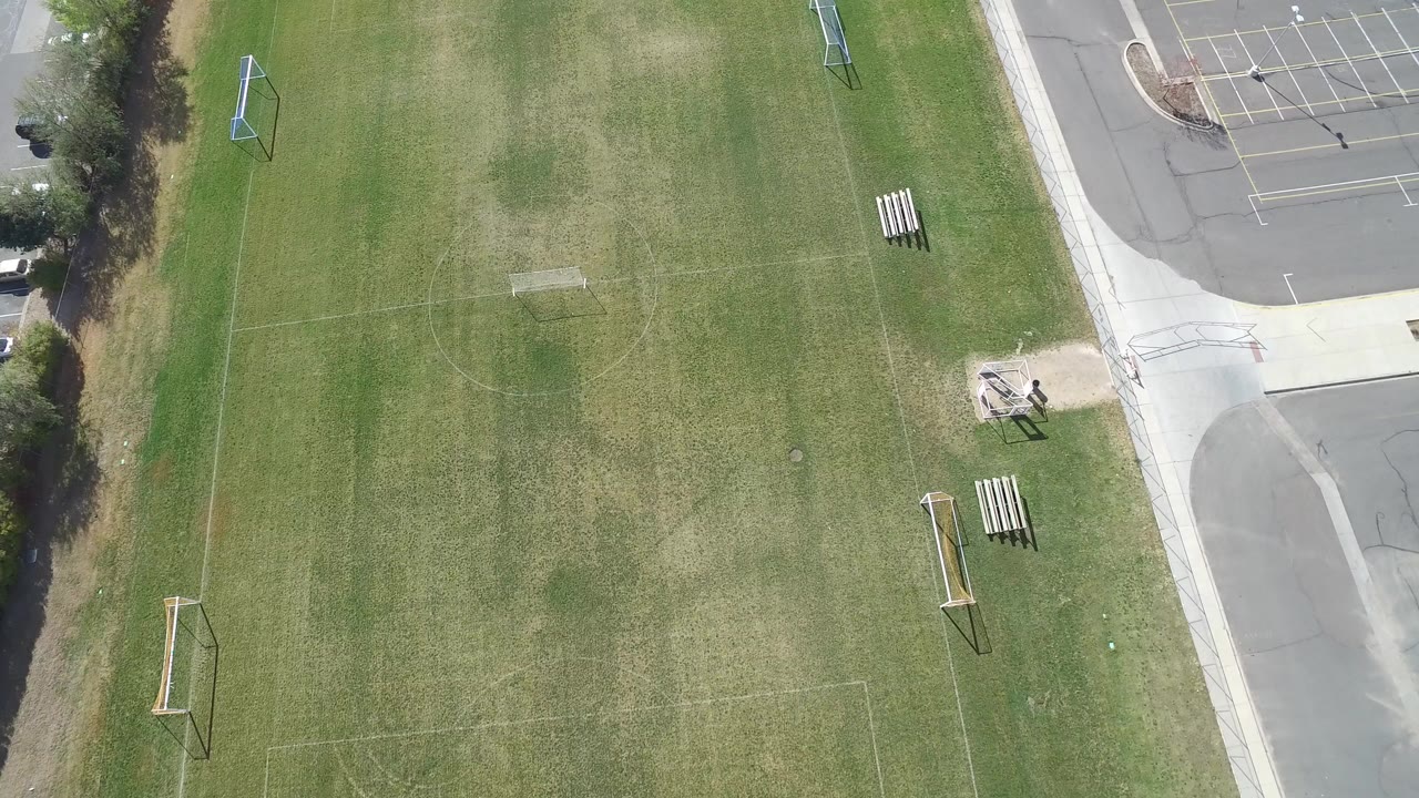 Overhead view of empty green sports fields beside a paved road and parking lot.