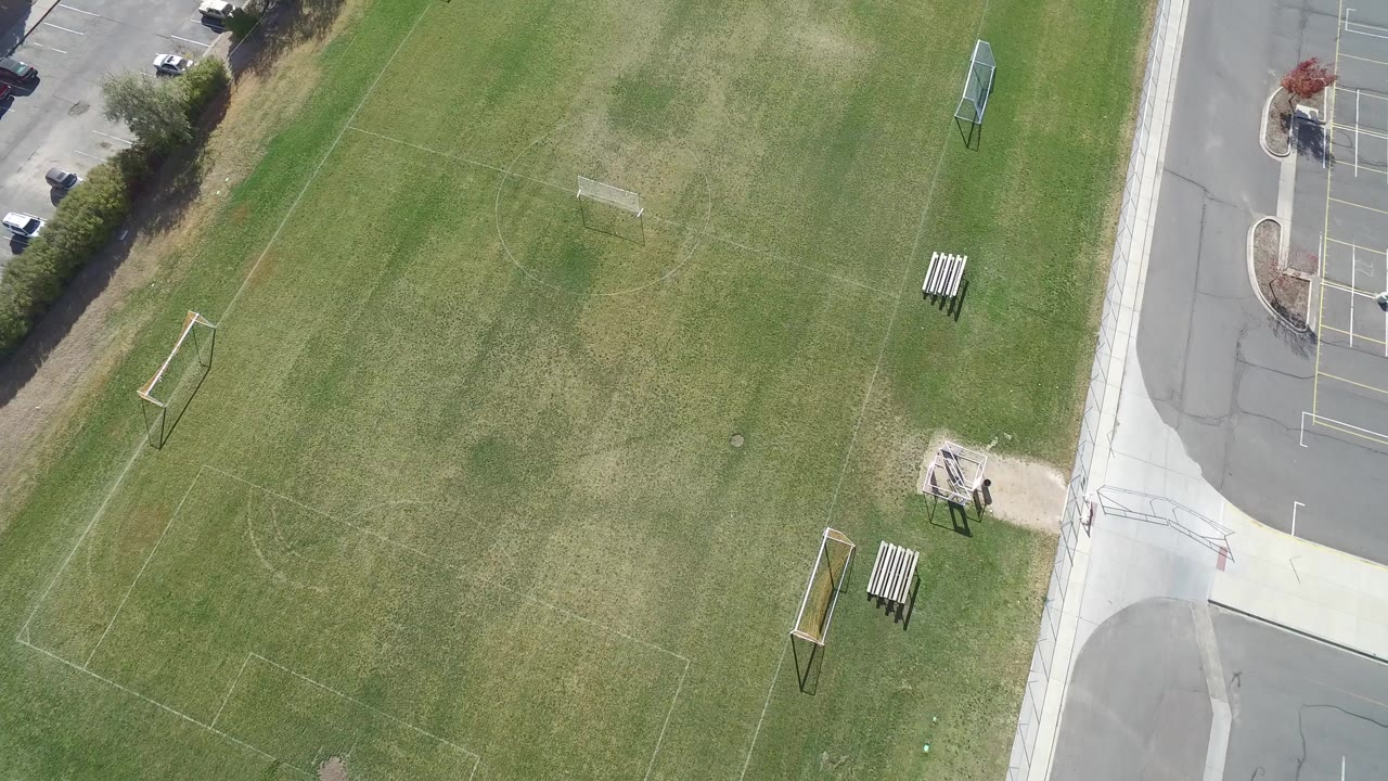 Aerial view over a Hidden Lake, Colorado soccer field with faint painted lines, portable goals, benches, a sidewalk, road edge, and no visible people.