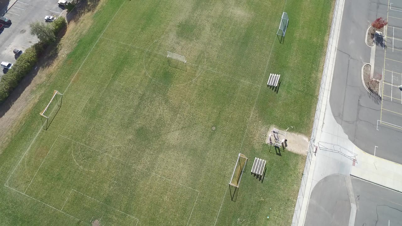 High drone frame of the Hidden Lake, Colorado field centered on worn green grass, faint soccer markings, two portable goals, a bench, road pavement, and no visible people.