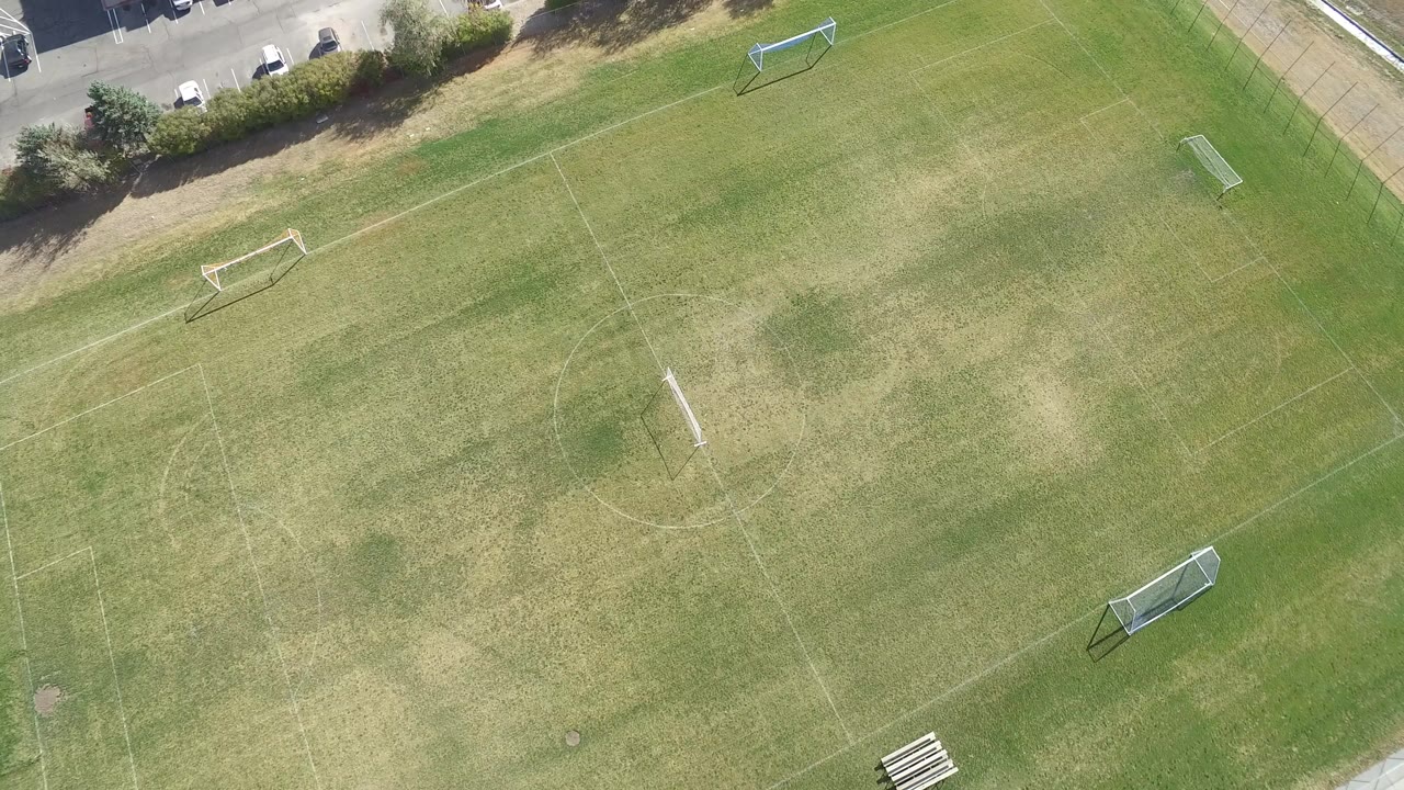 Overhead frame of an empty lined sports field in Hidden Lake, Colorado, with small goals, patchy grass, trees, and a parking lot edge, with no visible people.