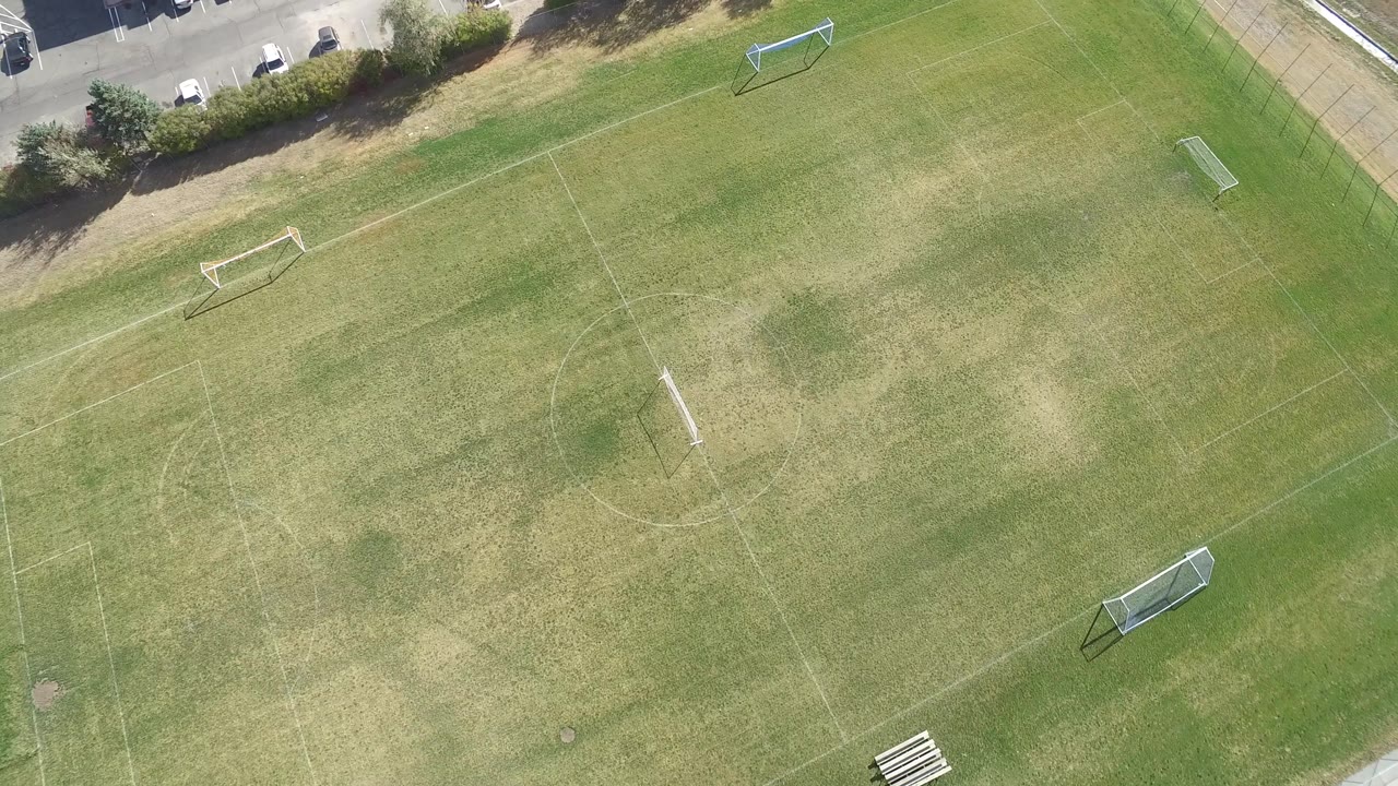 High overhead view in Hidden Lake, Colorado, showing the empty field centered around faint markings, movable goals, a paved path, and dry grass patches, with no visible people.