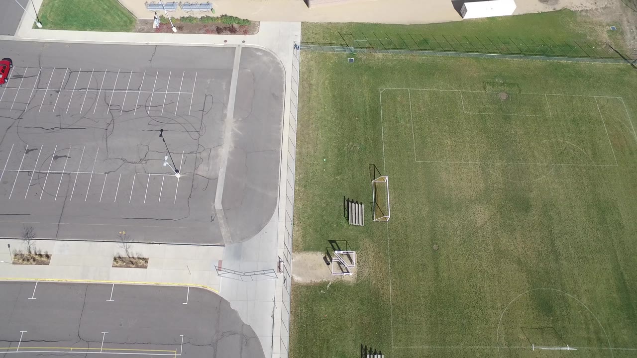 High aerial view in Hidden Lake, Colorado with empty parking rows on the left, a green field on the right, small bleachers near the edge, and no visible people.