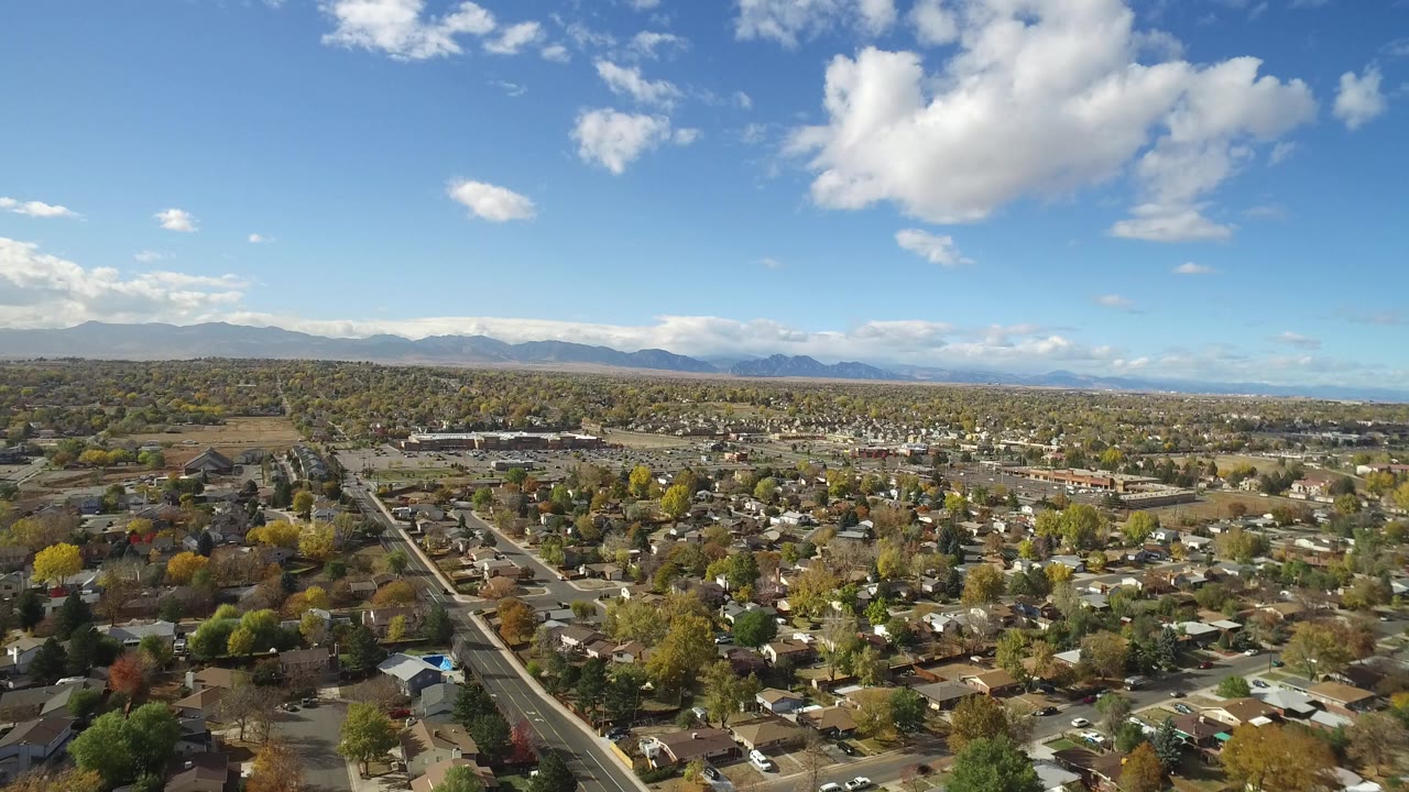 Opening aerial frame over Hidden Lake, Colorado, showing small homes, tree-lined streets, distant mountains, scattered clouds, and no visible people.