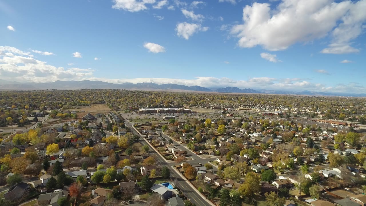 Early frame of Hidden Lake, Colorado showing a high aerial view over private homes, tree-lined streets, distant mountains, scattered clouds, and no visible people.