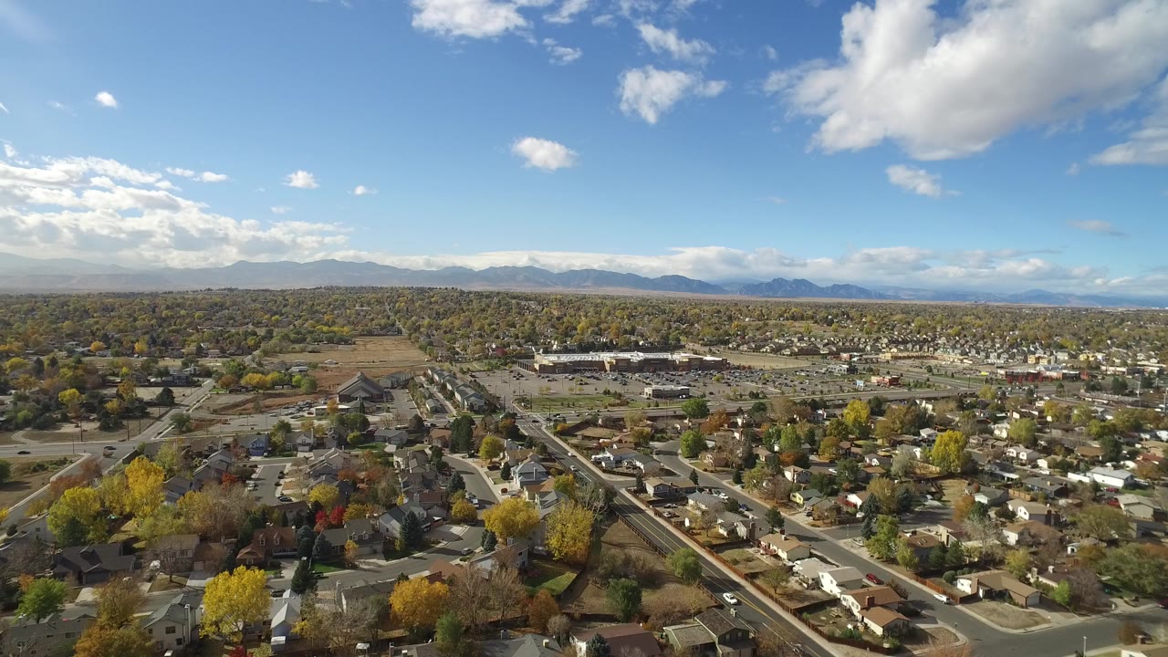 Aerial view of houses, streets, trees, and distant mountains under a cloudy blue sky.