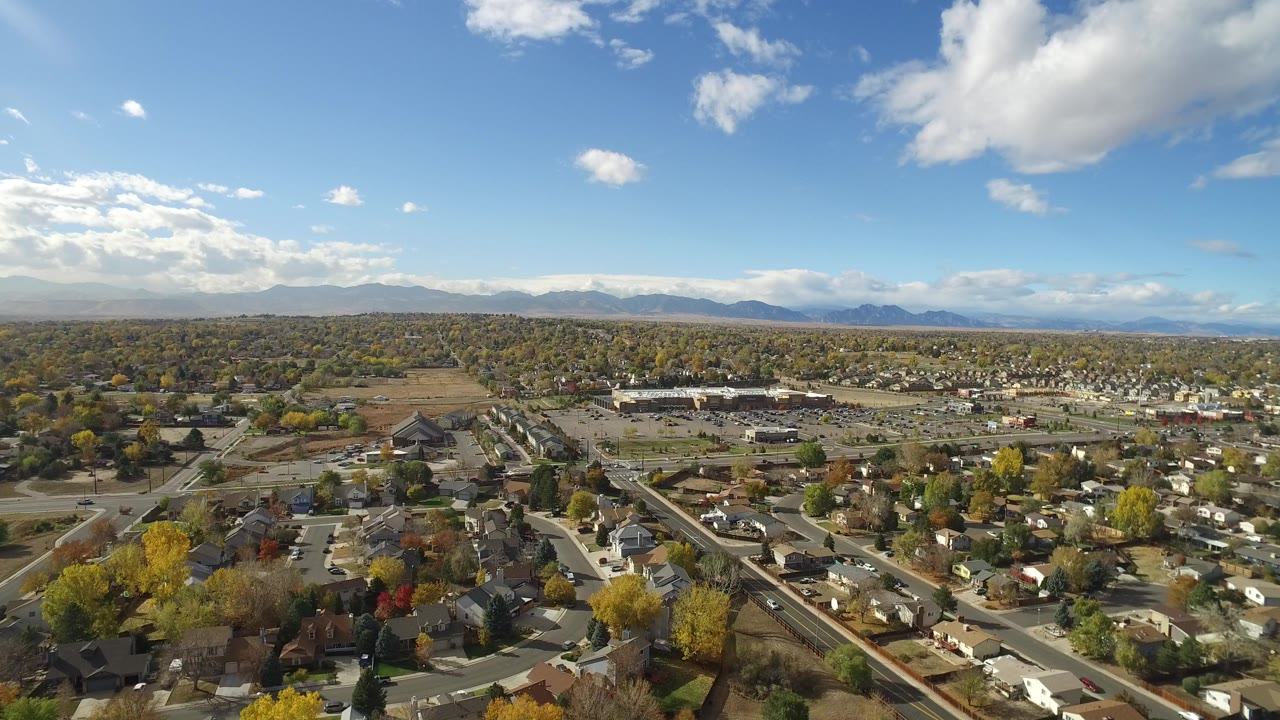 Wide aerial view over Hidden Lake, Colorado, with homes, autumn trees, open ground, distant mountains, scattered clouds, and no visible people.
