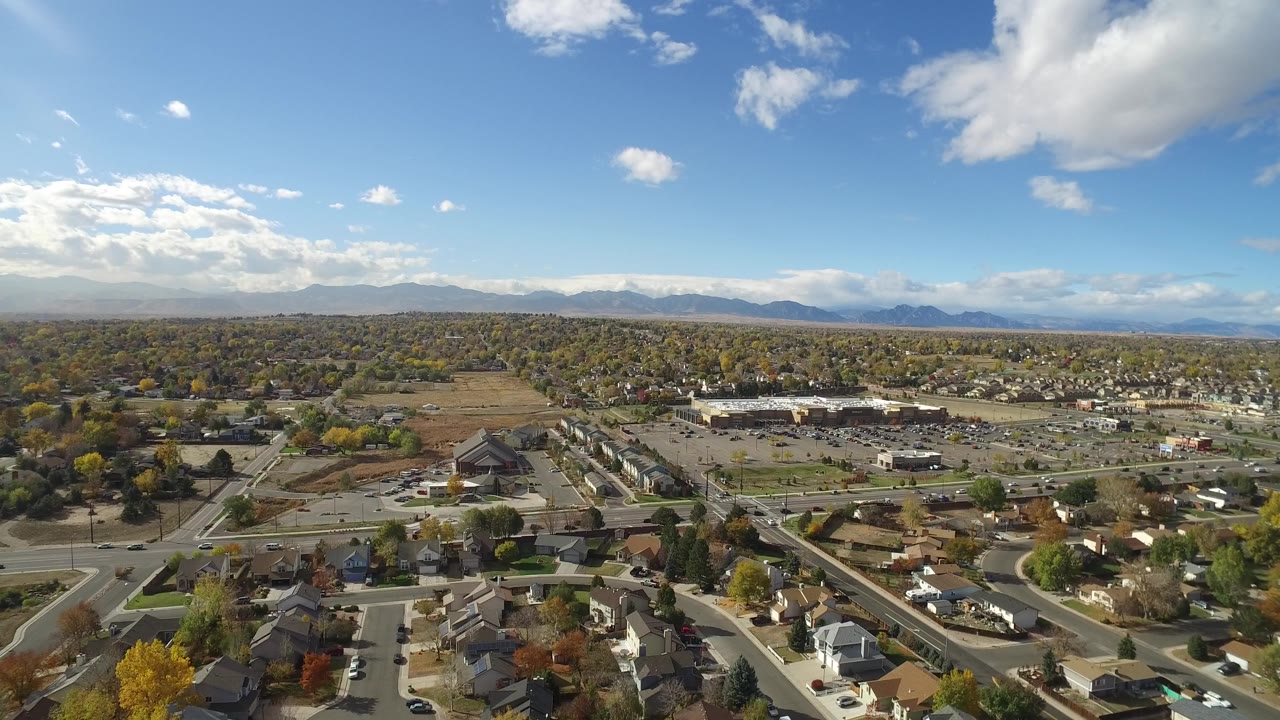 High aerial frame of Hidden Lake, Colorado showing rooftops and intersecting roads in the foreground, a large commercial parking area in the midground, mountains in the distance, and no visible people.