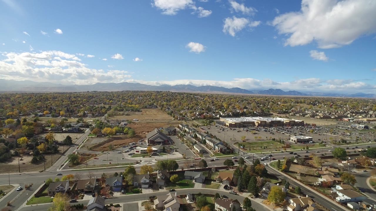 Wide aerial view of suburban streets, parking lots, trees, and distant mountains under blue sky.