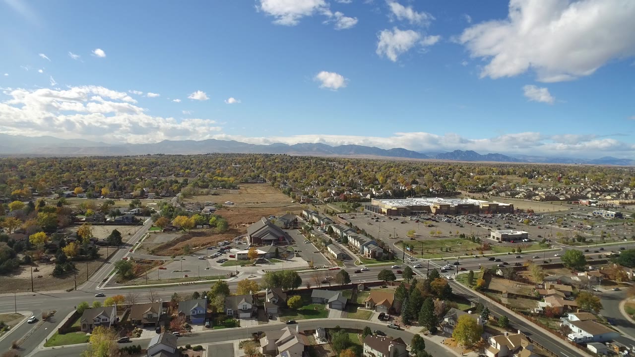 Aerial frame over Hidden Lake, Colorado, with rooftops along the lower edge, a main road crossing the scene, large parking lots, low buildings, autumn-colored trees, and no visible people.