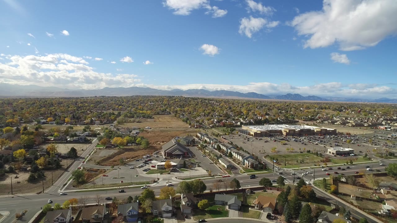 Second aerial frame over Hidden Lake, Colorado showing traffic on the foreground road, a large parking area, rows of homes, autumn trees, distant mountains, and no visible people.