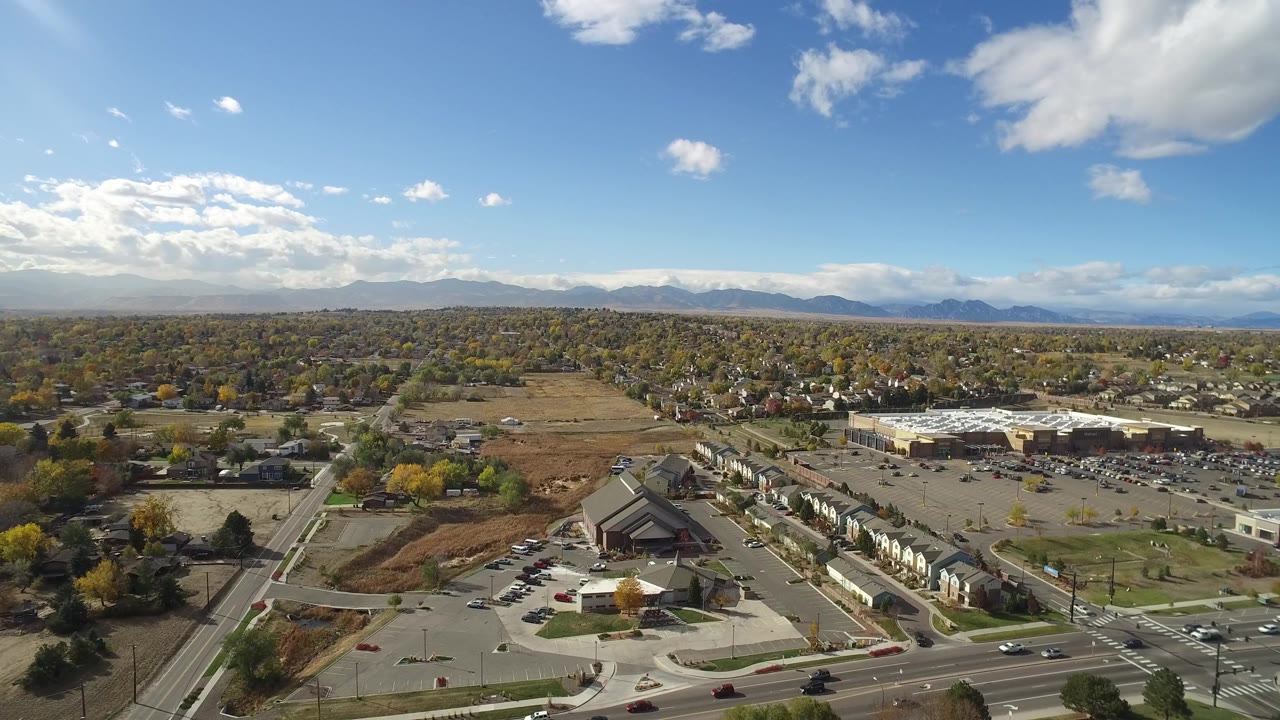 Opening aerial frame over Hidden Lake, Colorado with a broad road intersection, parking lots, homes, open fields, tree cover, distant mountains, bright clouds, and no visible people.