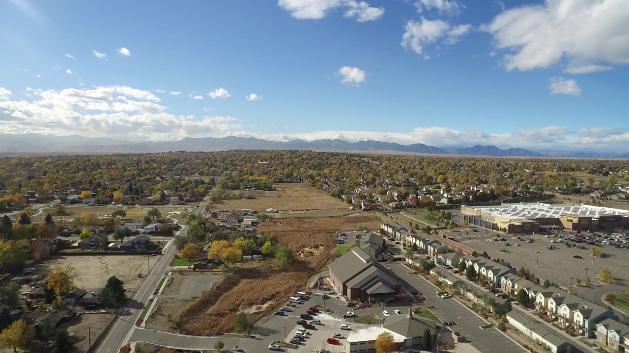 Aerial view over suburban streets, homes, parking lots, open land, and distant mountains.