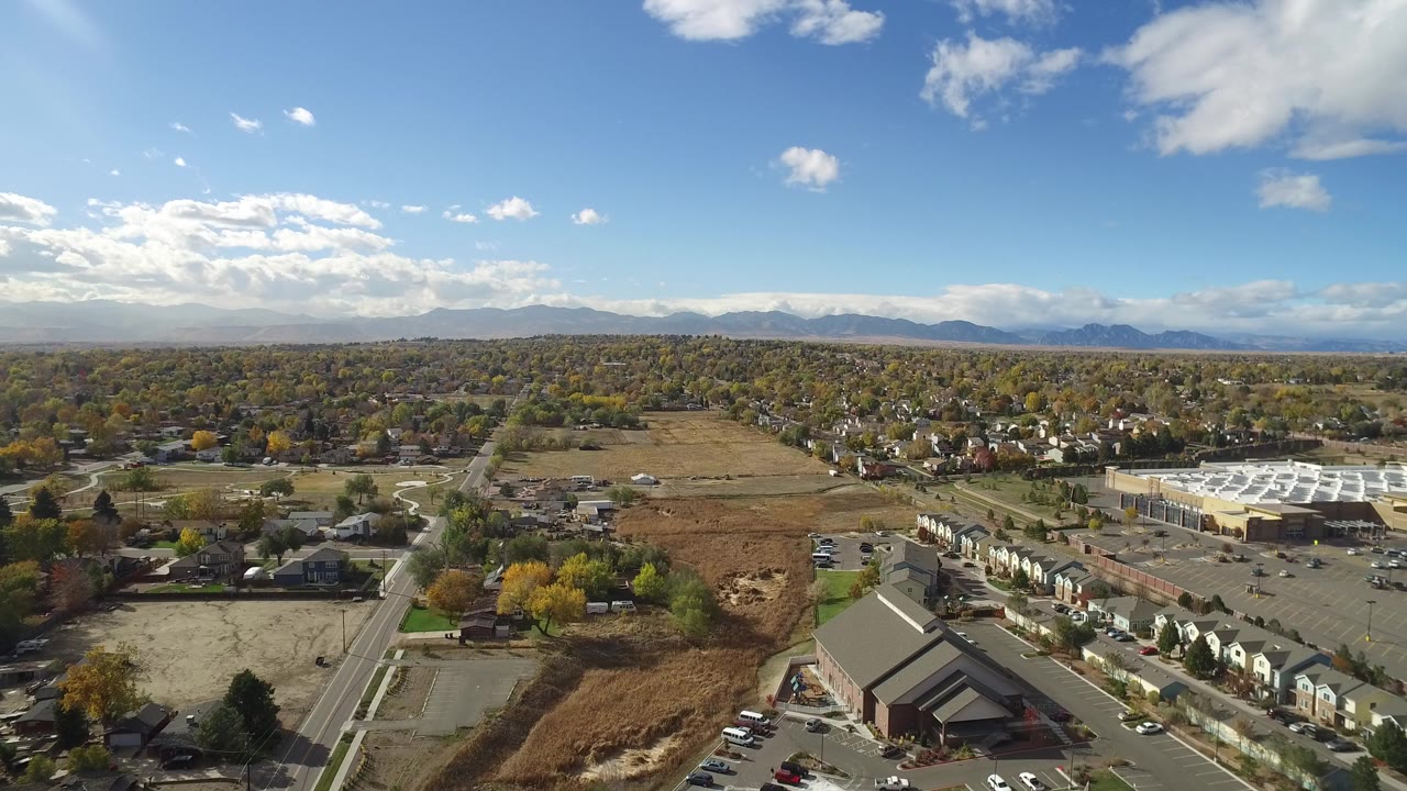 Drone-like view across homes, a large white-roofed building, roads, and fields.