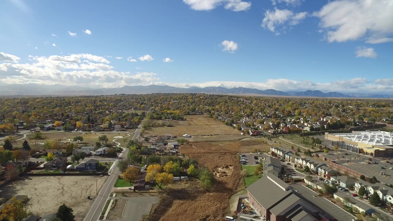 Aerial frame from Hidden Lake Colorado showing a road at left, scattered homes, brown open land, autumn-colored trees, distant mountains, and no visible people.