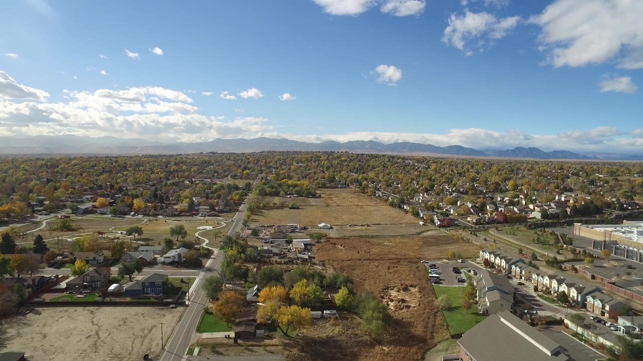 Opening aerial frame over Hidden Lake, Colorado with a road grid, homes, open brown fields, autumn trees, distant mountains, scattered clouds, and no visible people.