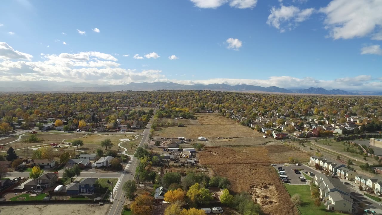 Aerial suburban landscape with a large open field, houses, roads, trees, and a clouded mountain horizon.
