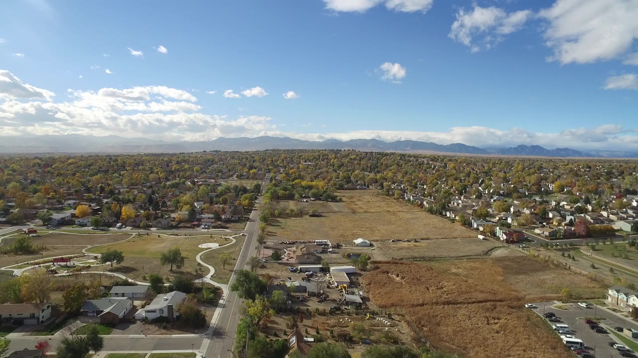 Aerial view over suburban streets, open fields, houses, trees, and distant mountains.