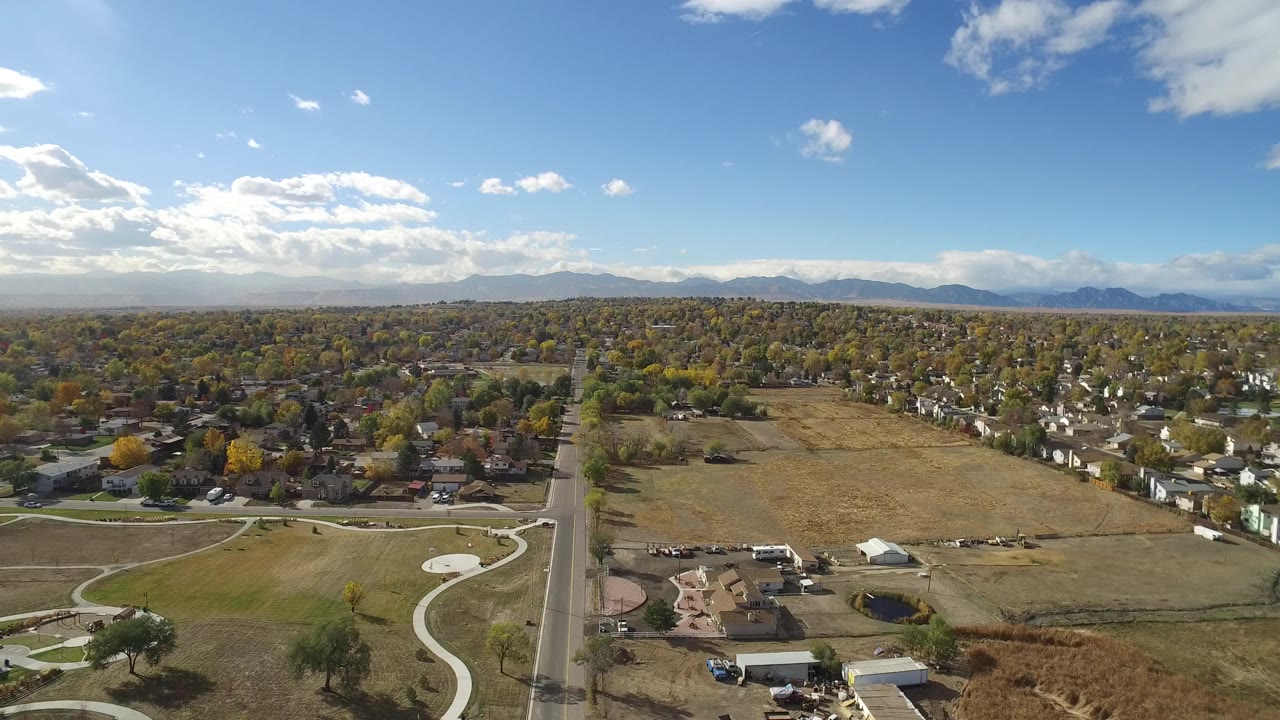 Wide aerial frame in Hidden Lake, Colorado showing residential blocks, a large tan field, curving paved paths in the foreground, distant mountains, and no visible people.
