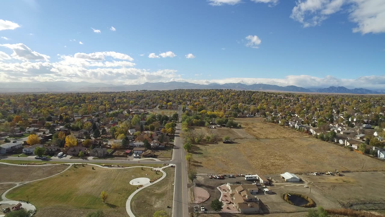 Opening aerial frame over Hidden Lake, Colorado, showing a straight road, curving park paths, dry open land, residential streets, distant mountains, and no visible people.