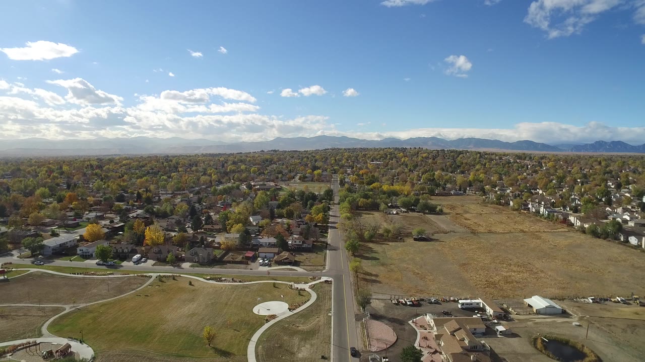 Opening aerial frame over Hidden Lake, Colorado, with a straight road dividing lawns and homes, autumn trees across the neighborhood, distant mountains, scattered clouds, and no clearly visible people.