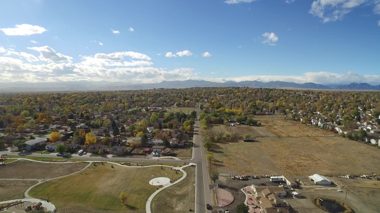 Aerial frame in Hidden Lake, Colorado, showing the road centered below, a broad tan open lot to the right, houses and trees beyond it, distant mountains, and no clearly visible people.
