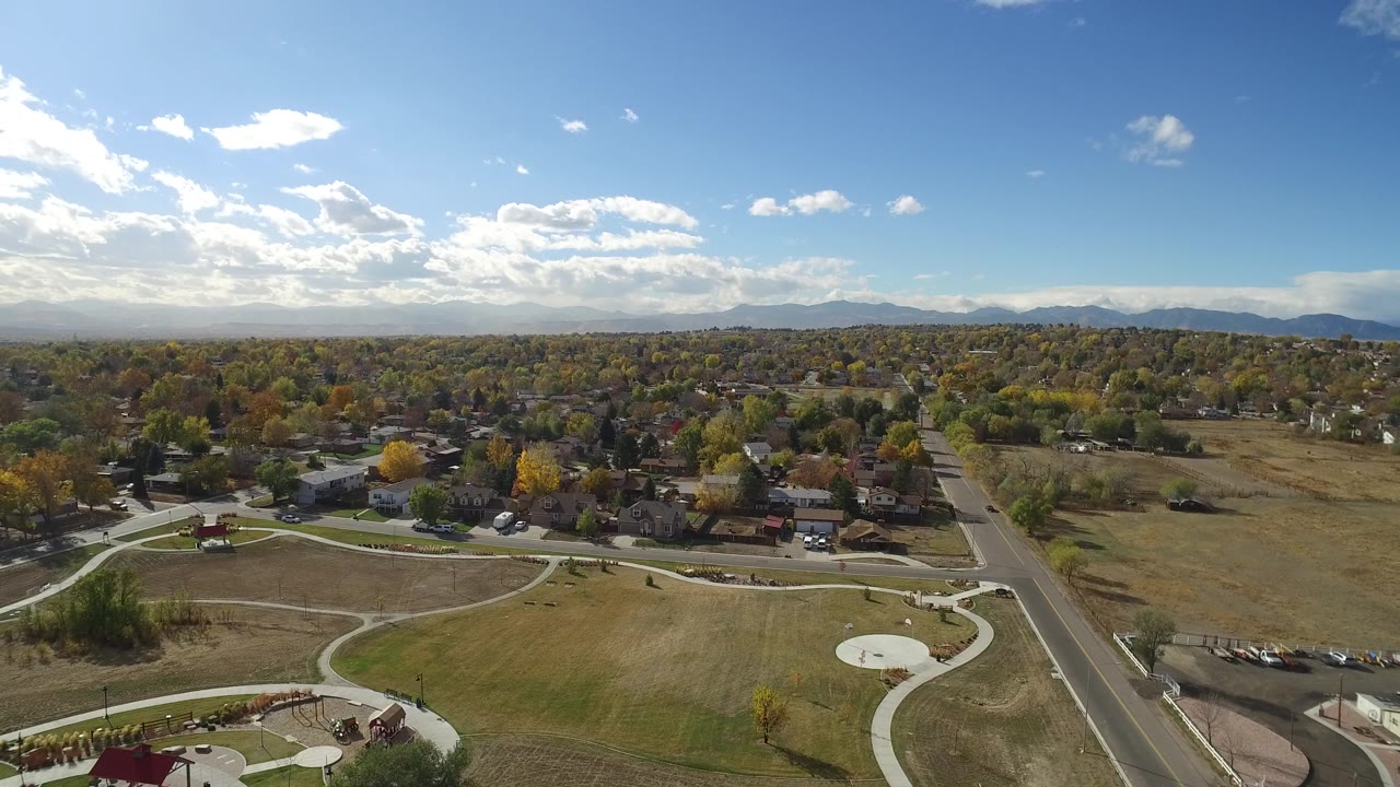 Second aerial frame in Hidden Lake, Colorado showing the road more centrally, open fields to the right, houses beyond the park, distant mountains, and no visible people.