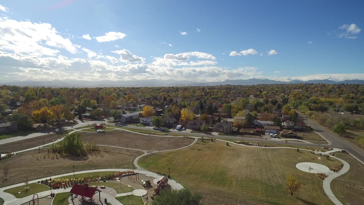 Opening aerial frame over Hidden Lake, Colorado, showing a grassy park with curved paths, a playground roof at the bottom edge, residential streets, tree cover, distant mountains, and no visible people.