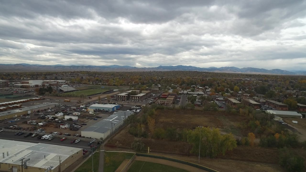 High aerial view over Hidden Lake, Colorado with white-roofed industrial buildings, parked cars, autumn trees, distant neighborhoods, mountains, and no visible people.
