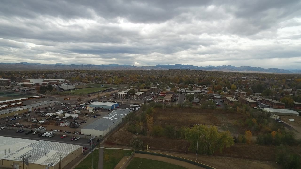 Drone-height view across Hidden Lake, Colorado showing commercial rooftops, an open brown field, parked vehicles, roads, tree lines, distant homes, mountains, and no visible people.
