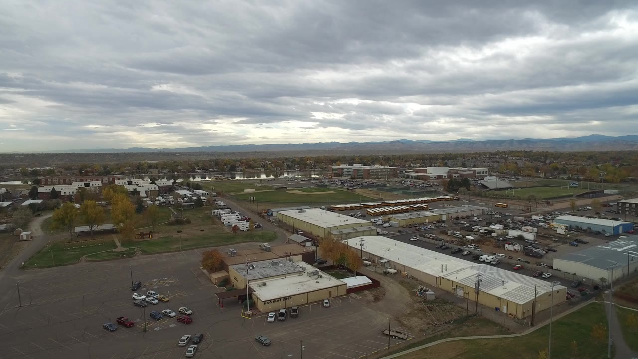 High aerial frame over Hidden Lake, Colorado, showing low commercial buildings, parking areas, distant neighborhoods, mountains, and no visible people.