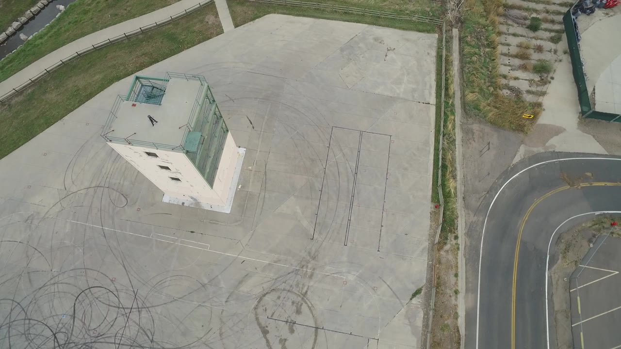 Top-down aerial frame in Hidden Lake, Colorado showing a pale rectangular building, concrete pad, curving road edge, grass strips, and dark tire marks with no visible people.