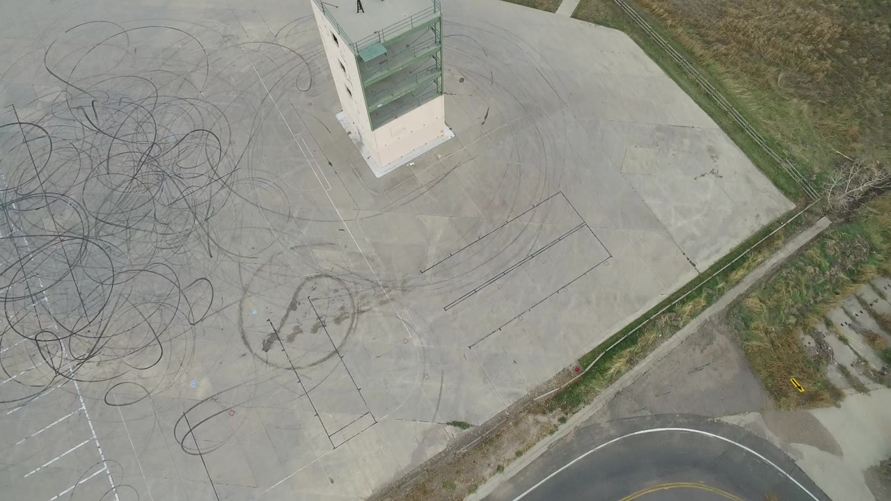 Overhead frame of an abandoned parking lot in Hidden Lake, Colorado, showing looping tire marks, faded pavement markings, grass borders, and no visible people.