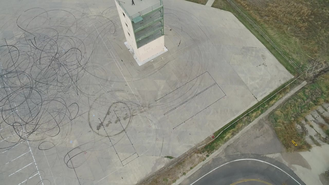Drone-style overhead frame in Hidden Lake, Colorado, with the rectangular structure near the top, dense tire scuffs across the lot, parking stripes, grass, and no visible people.