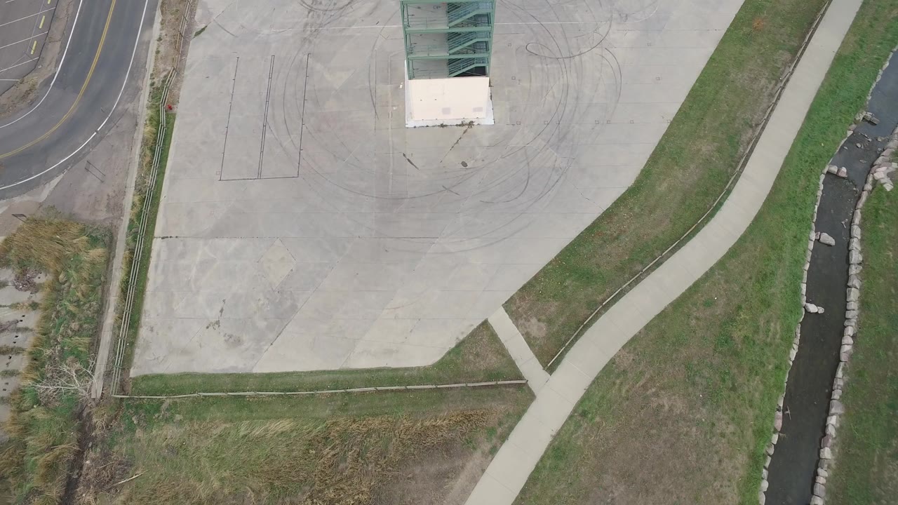 Overhead view of concrete plaza, curving road, sidewalk, grass, and drainage channel.