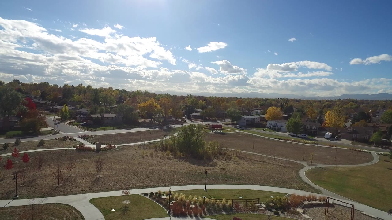Opening aerial view in Hidden Lake, Colorado, showing curving park paths, dry grass, nearby homes, autumn trees, distant mountains, bright sun, and no visible people.